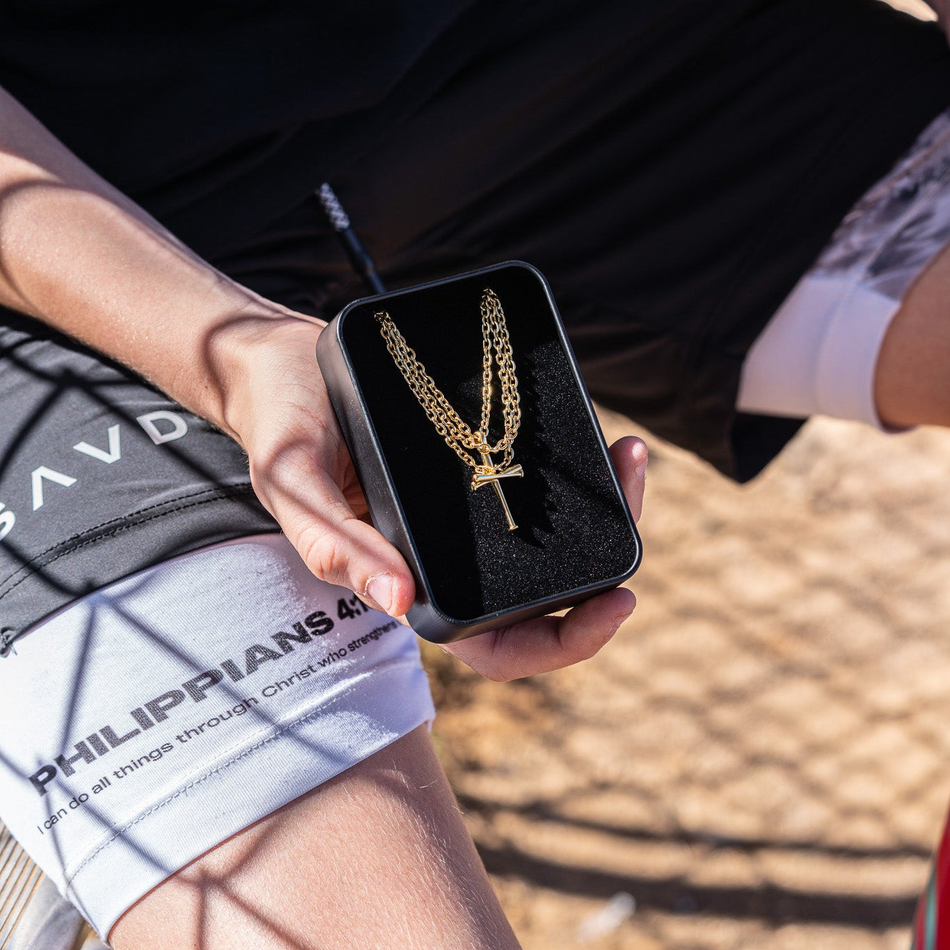Christian Baseball Bat Cross Necklace: Person holding a gold baseball bat cross necklace in a black box with a blurred background