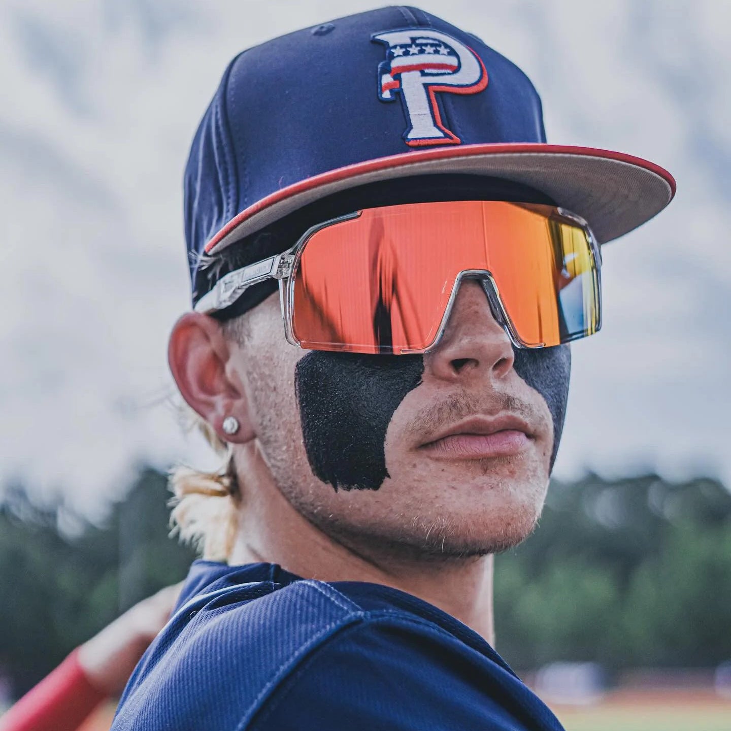 Baseball Sunglasses | A baseball player in a navy blue uniform and matching cap with a red brim, wearing reflective orange-tinted shield sunglasses and black face paint, photographed outdoors on a field.
