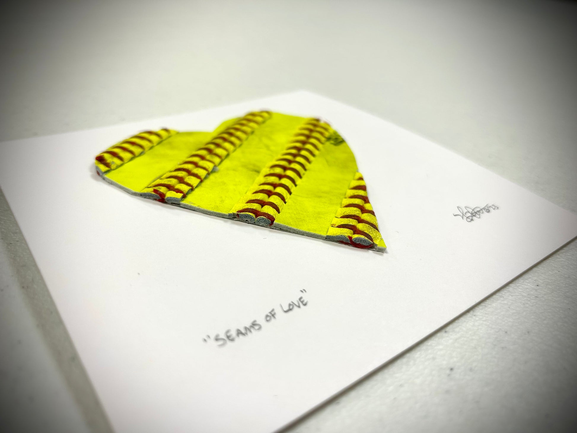 Softball Heart Artwork titled “Seams of Love” with yellow leather seams on a heart shaped design.