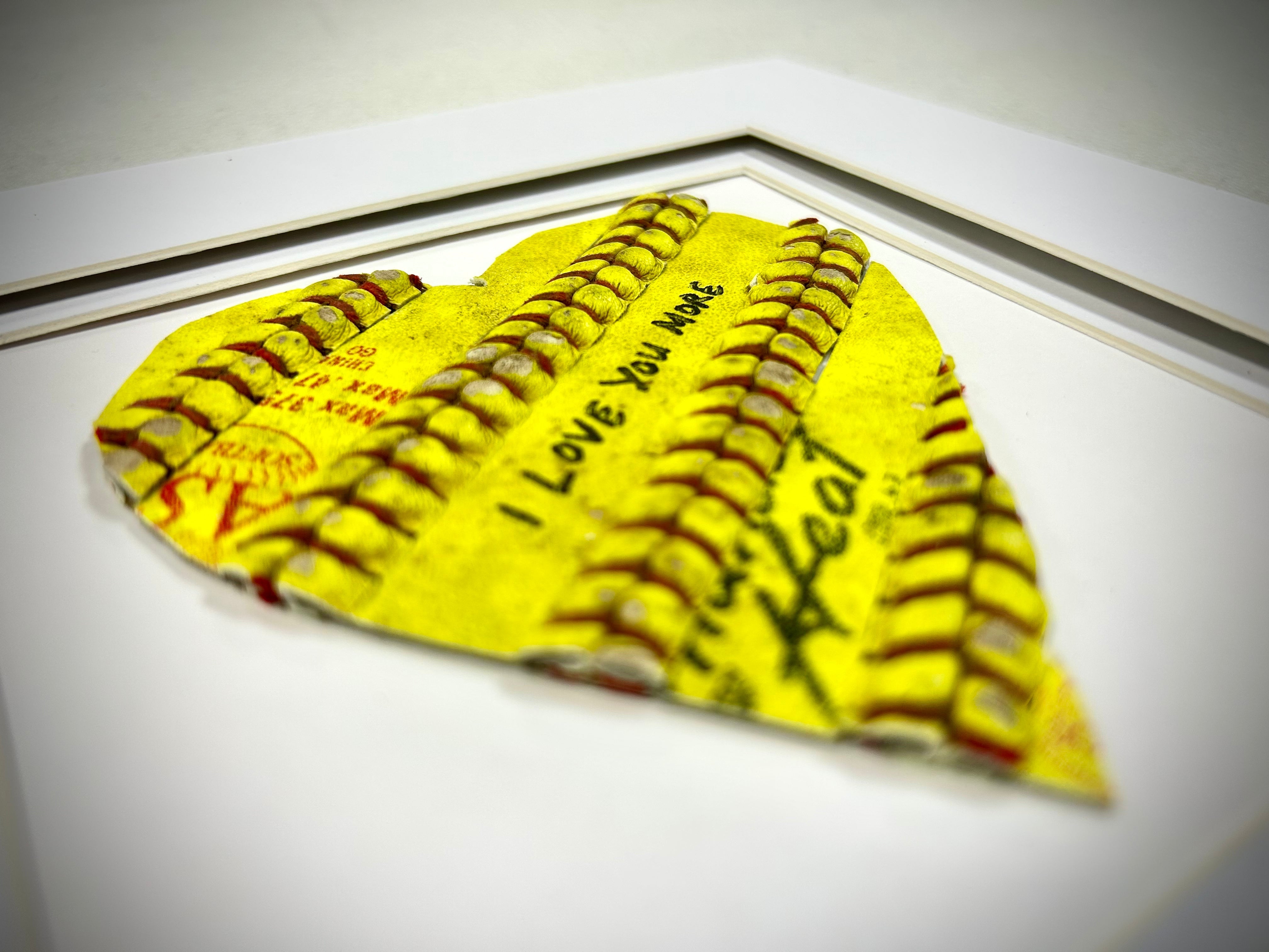 Softball Heart Artwork titled “Seams of Love” featuring a handcrafted heart with yellow leather and red stitching.