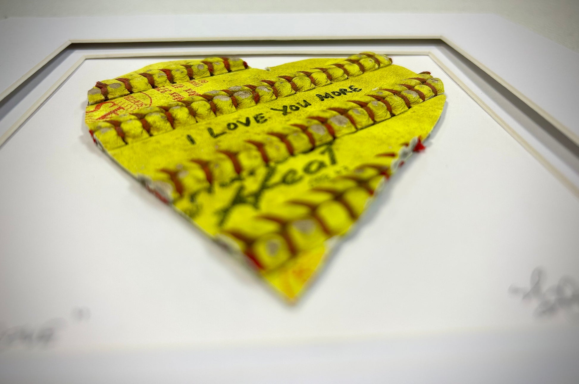 Softball Heart Artwork titled “Seams of Love” showcasing a heart-shaped piece with the phrase 'I LOVE YOU MORE'.