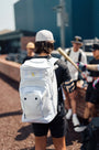 Luccini Baseball Wheeled Gear Bag – White | Baseball player wearing a white backpack with a blurred background of people and a building.