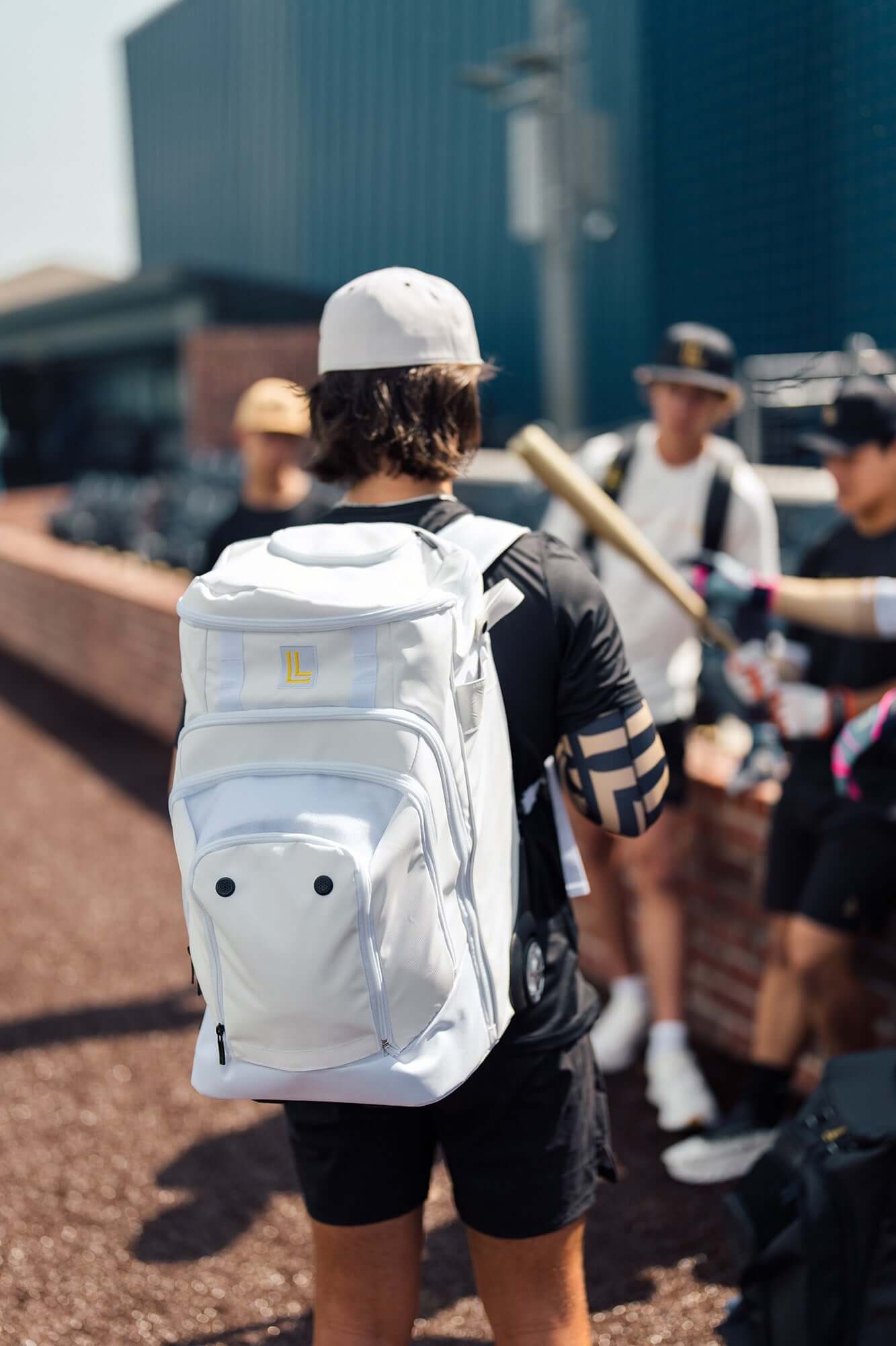 Luccini Baseball Wheeled Gear Bag – White | Baseball player wearing a white backpack with a blurred background of people and a building.