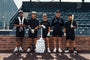 Luccini Baseball Wheeled Gear Bag – White | Five baseball players in black uniforms standing in front of a brick wall with stadium seats in the background.