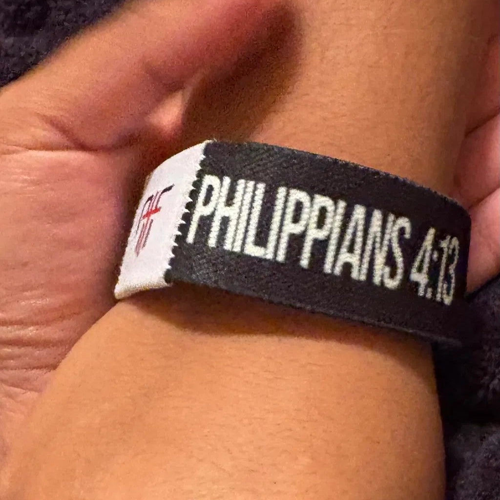 Bible Verse Bracelet | A black wristband with white text reading 'PHILIPPIANS 4:13' worn on a person's wrist, with a hand holding the wristband in place against a dark textured background.