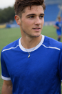 Man wearing a blue sports jersey on a grassy field with a soccer cross necklace