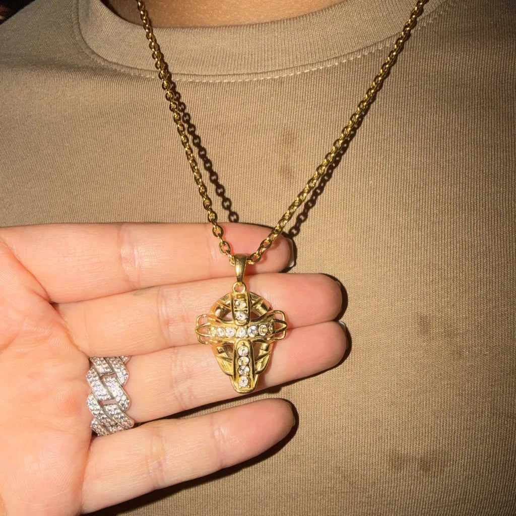 Iced Out Baseball Catcher Mask Necklace | A gold cross pendant necklace with clear stones, worn on a chain and held in a hand against a beige t-shirt background.